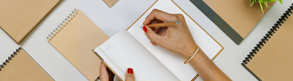 A hand holding open a notebook and another writing within the notebook, surrounded by a variety of tan notebooks.