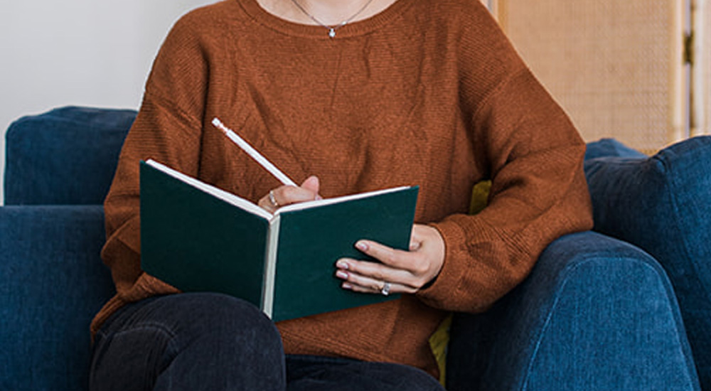 A woman writing down in an open journal sitting on a couch facing the camera.