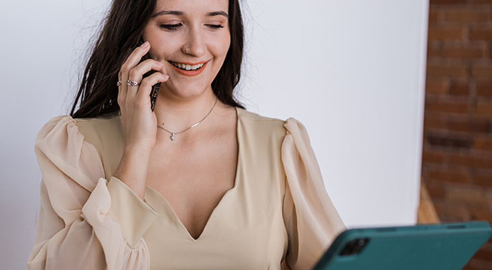 A woman talking on the phone and smiling down at her tablet while standing in front of the camera.